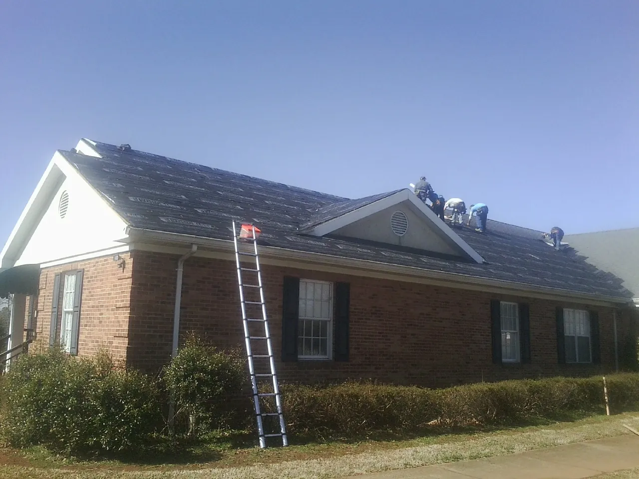 Crew tearing shingles off a brick ranch roof with a ladder set — Emergency Roof Repair work by our crew in Gonzales
